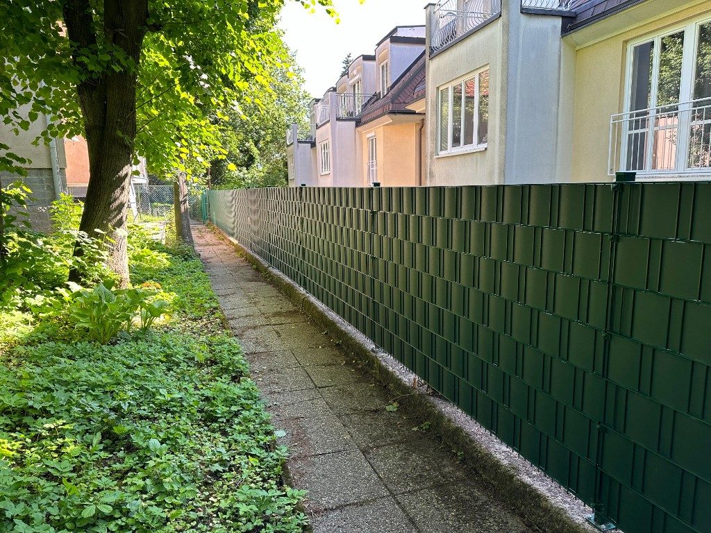 Dunkelgrüner Stabmattenzaun entlang eines Gehwegs vor Wohnhäusern